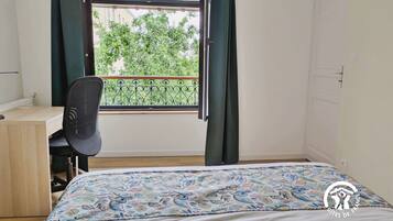 A bedroom with a bed, a desk, and a chair. There is a window with curtains and a view of greenery outside.