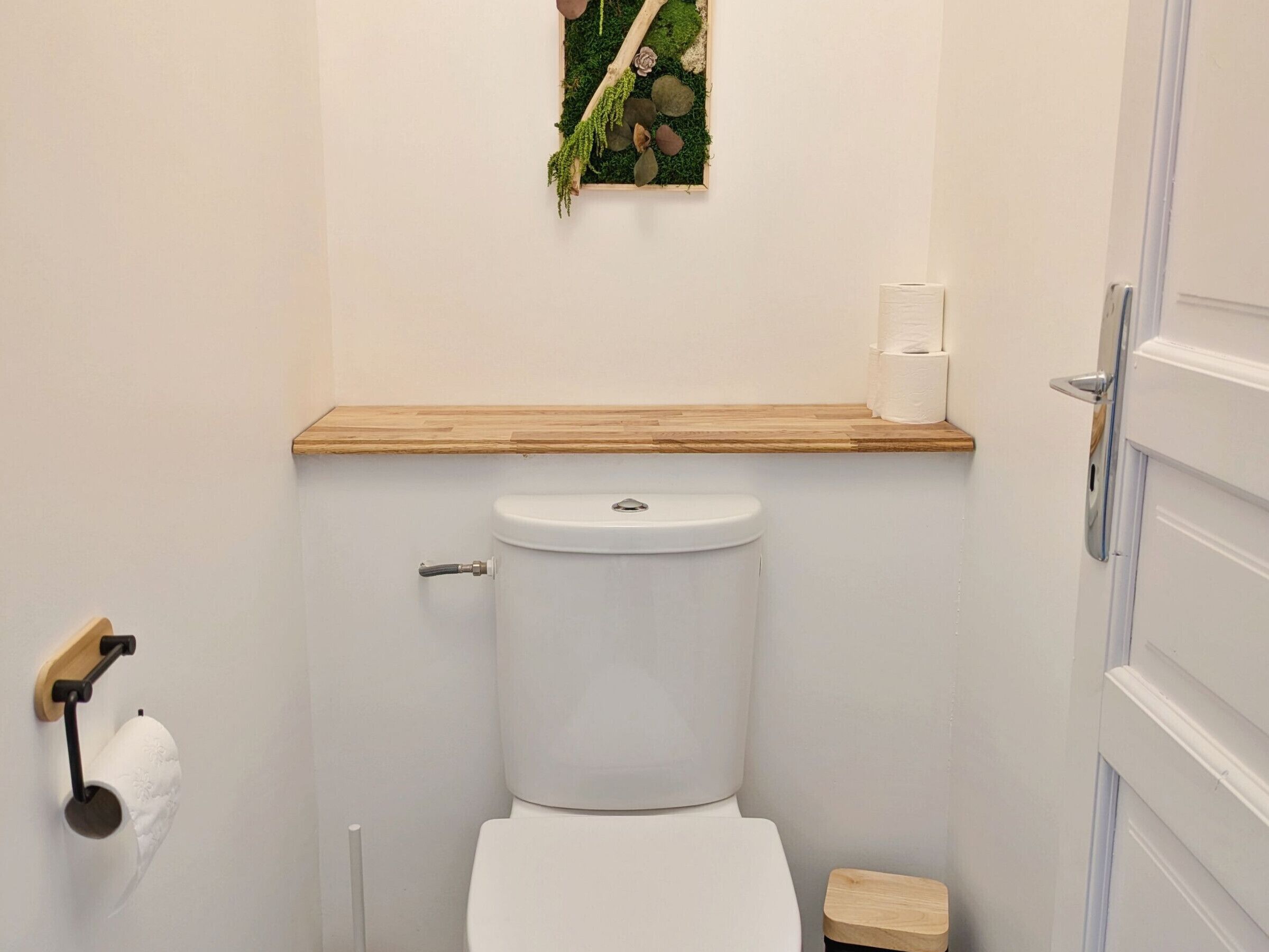 A small bathroom with a toilet, a wooden shelf above the toilet, a toilet paper holder, and a small wooden stool.