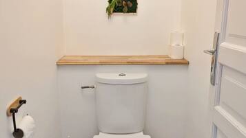 A small bathroom with a toilet, a wooden shelf above the toilet, a toilet paper holder, and a small wooden stool.