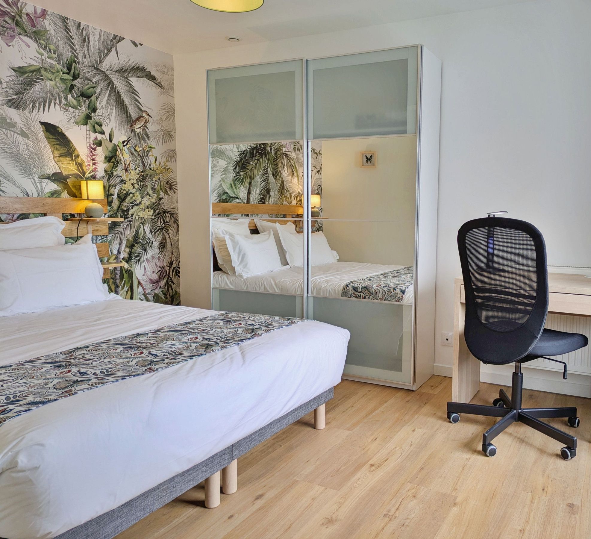 Ein Schlafzimmer mit einem Bett, einem Schreibtisch und einem Stuhl. Es gibt einen Kleiderschrank mit Glastüren und tropischem Wandpapier.