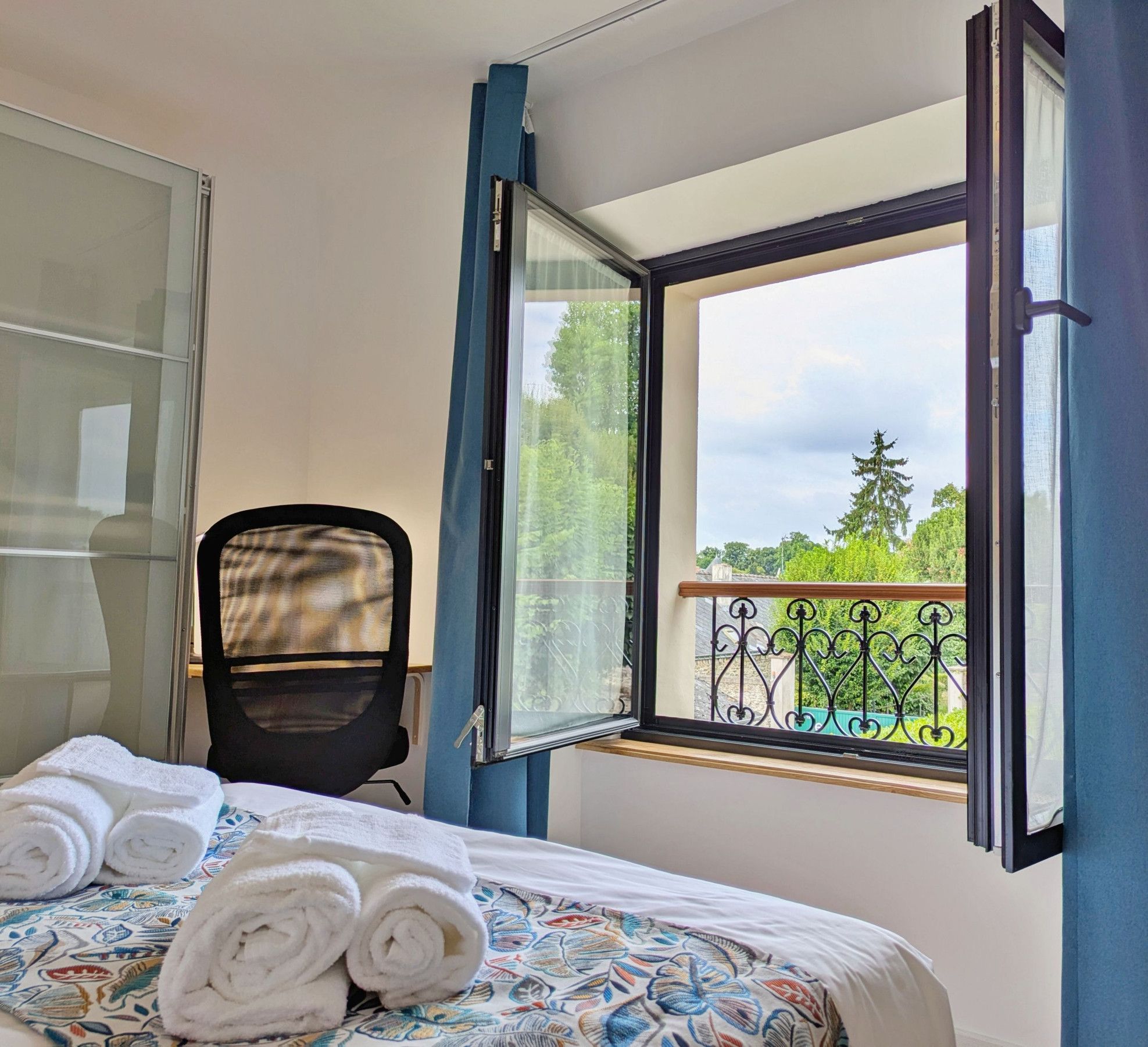 A hotel room with a bed, a chair, a window with curtains, and a view of greenery outside.