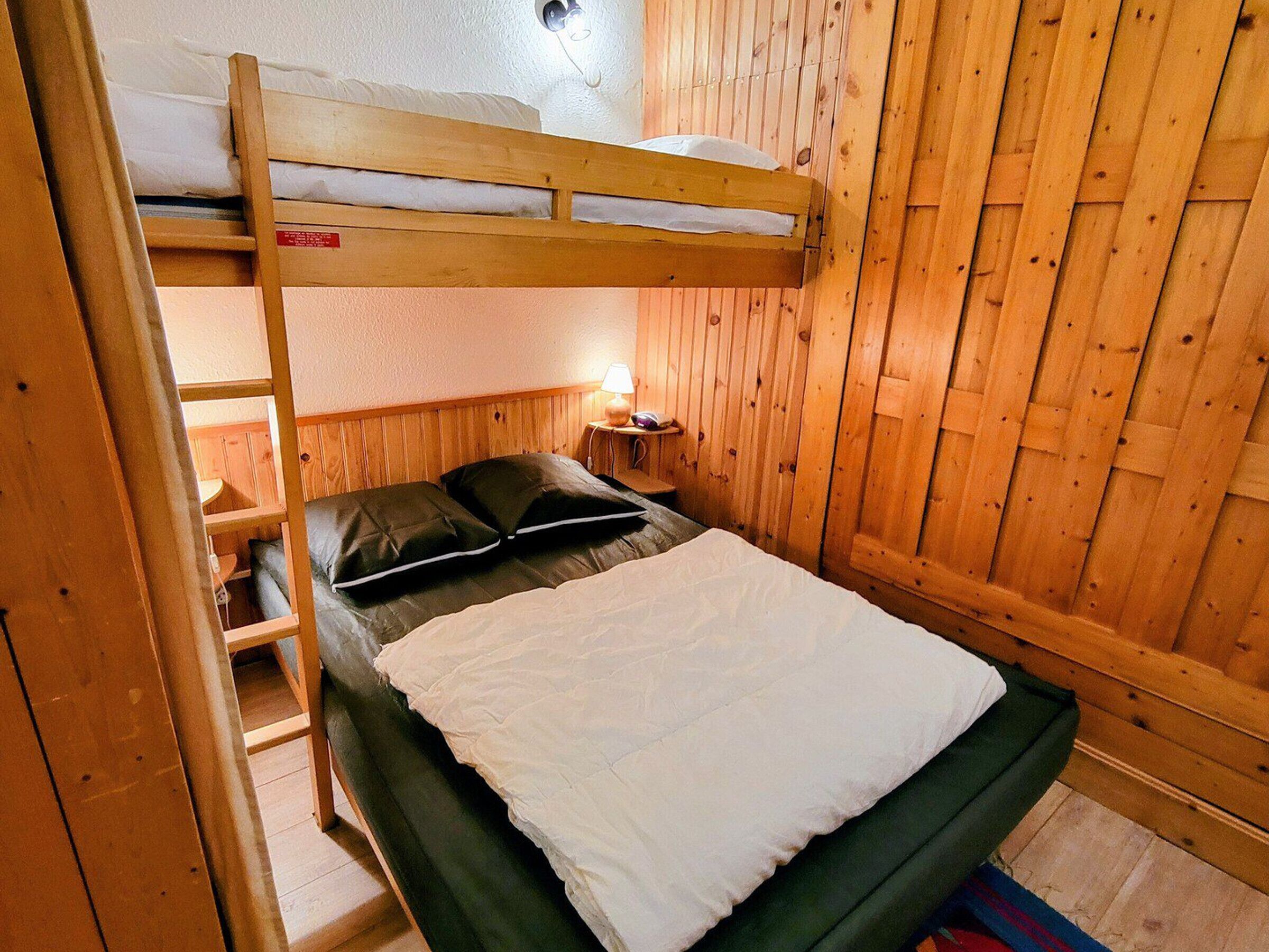 Una cama litera con cabecera de madera, una mesita de noche con lámpara y una ventana con persianas de madera.