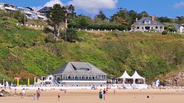 Ein Strand mit einem Gebäude und einem großen Balkon mit Blick aufs Meer, einem weißen Zelt und Personen im Sand.