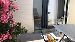 Un patio con una mesa y sillas, una puerta corrediza de vidrio que da a un espacio interior, y una maceta con flores rosas.