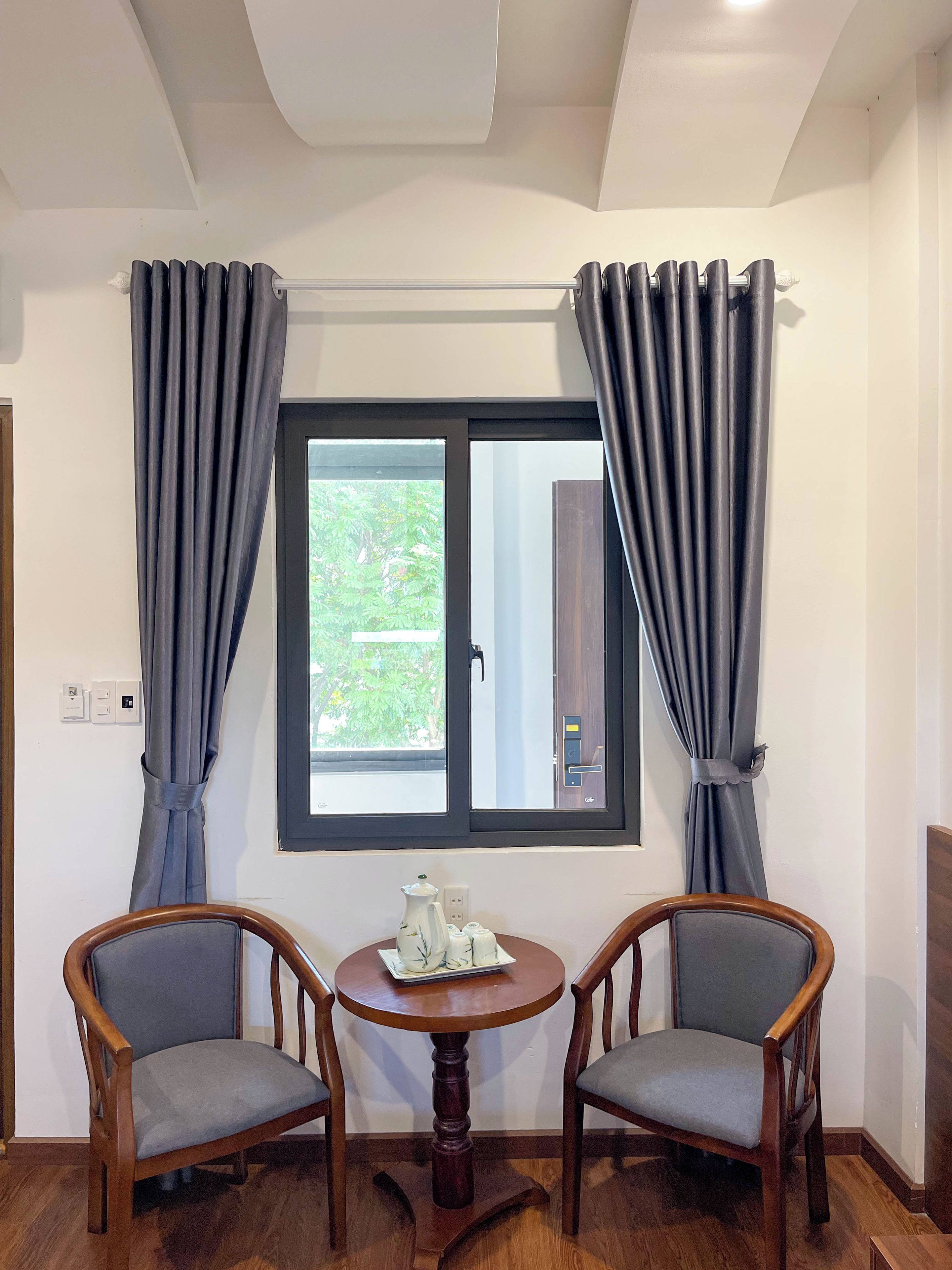 Una habitación con dos sillas de madera, una mesa redonda pequeña, una ventana con cortinas y una puerta con pomo.