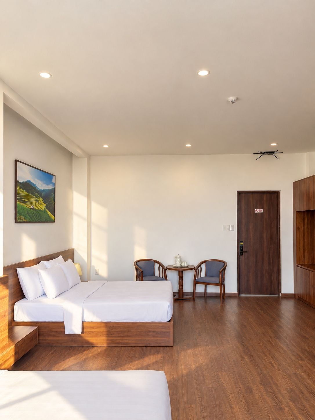 A hotel room with a bed, two chairs, a small table, a door, and a painting on the wall.