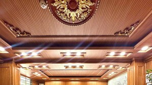 Eine Hotelhalle mit einer Rezeption, drei Stühlen, einem Marmortisch mit einer Karaffe und Obst, sowie einer Decke mit aufwendigen Holzschnitzereien und Beleuchtung.