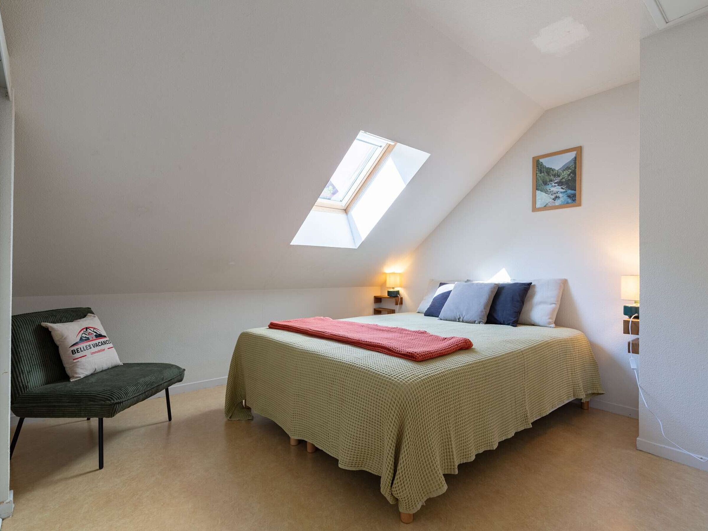 Une chambre avec un plafond en pente, un puits de lumière, un lit avec des oreillers, une chaise et une table de chevet avec une lampe.