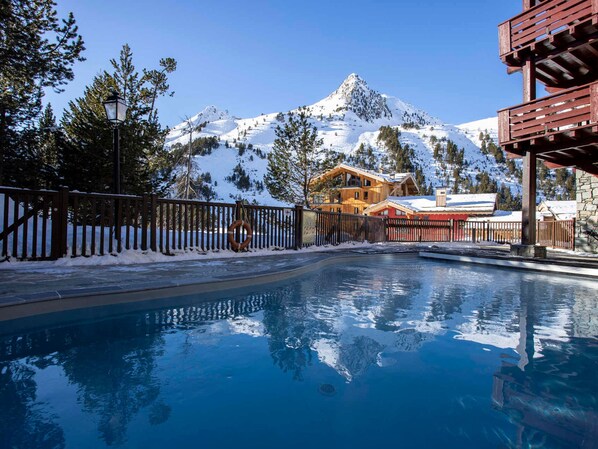 A swimming pool with a mountain backdrop, wooden buildings, and a snow-covered landscape.