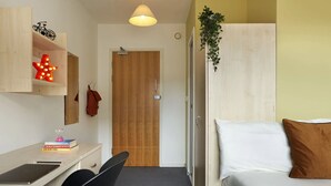 A compact room with a bed, a desk, and a chair. There is a wooden door, a hanging plant, and a wall-mounted shelf with a bicycle and a star-shaped light. - Students Only - Central London Student Accommodation (London)