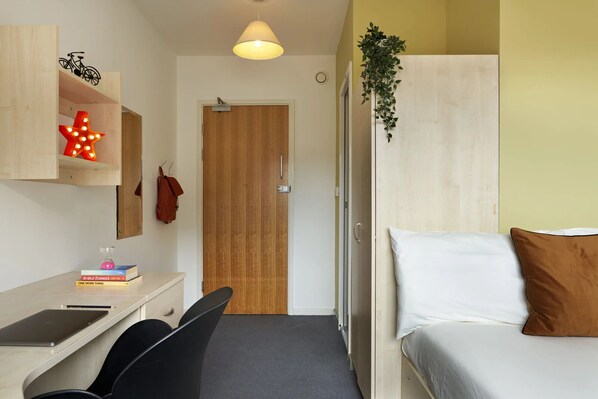 A compact room with a bed, a desk with a chair, a hanging plant, and a door. - Students Only - Central London Student Accommodation (London)