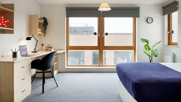 A bedroom with a bed, a desk with a chair, a window with blinds, a clock, and a radiator. - Students Only - Central London Student Accommodation (London)