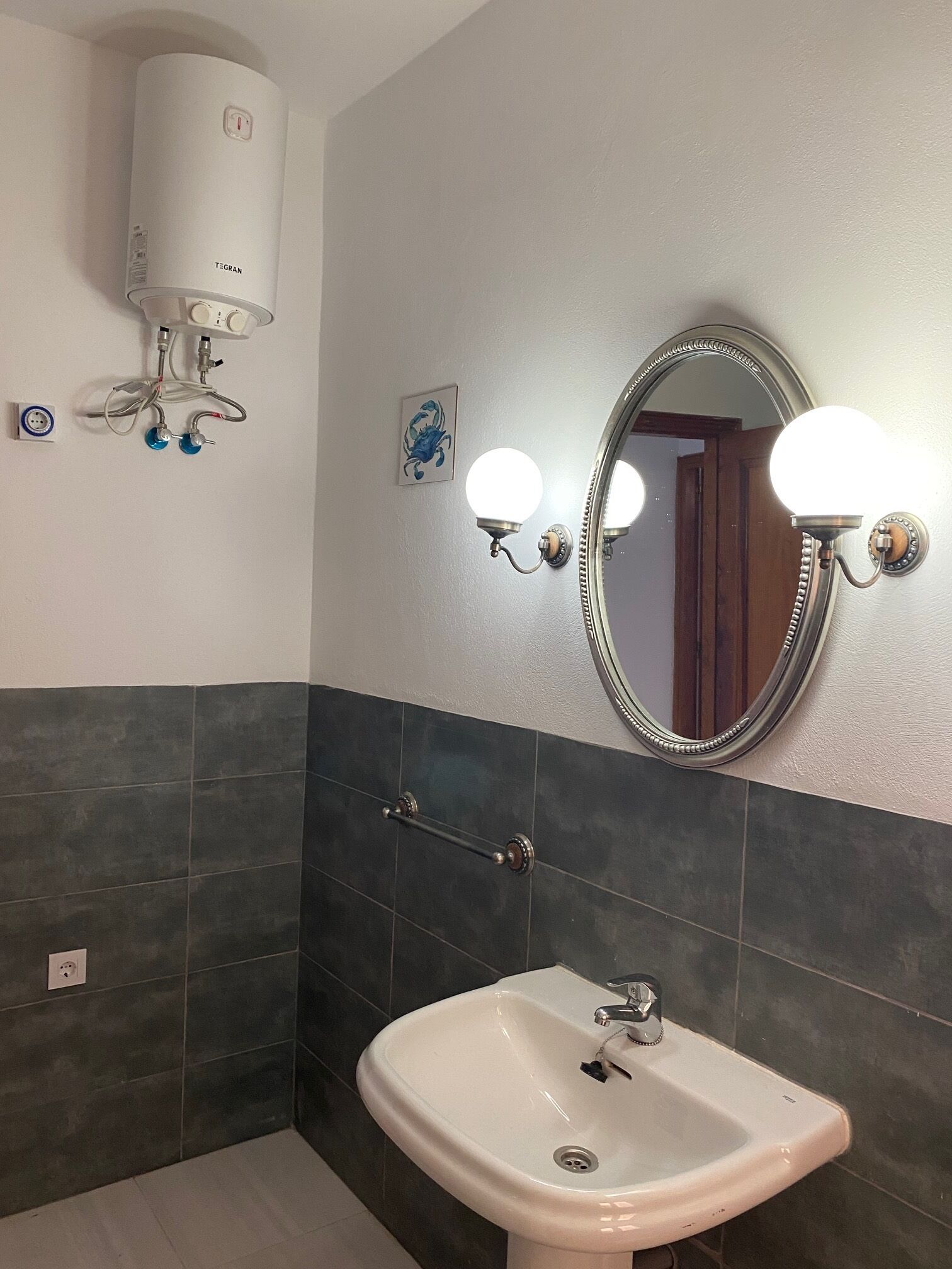 Ein Badezimmer mit einem weißen Waschbecken, dunkel gefliesten Wänden, einem Spiegel mit Beleuchtung und einem Handtuchhalter.