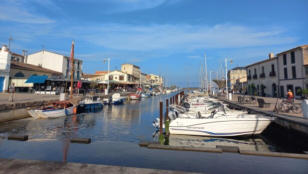 A marina with boats docked, a waterfront promenade, and buildings with shops and cafes.