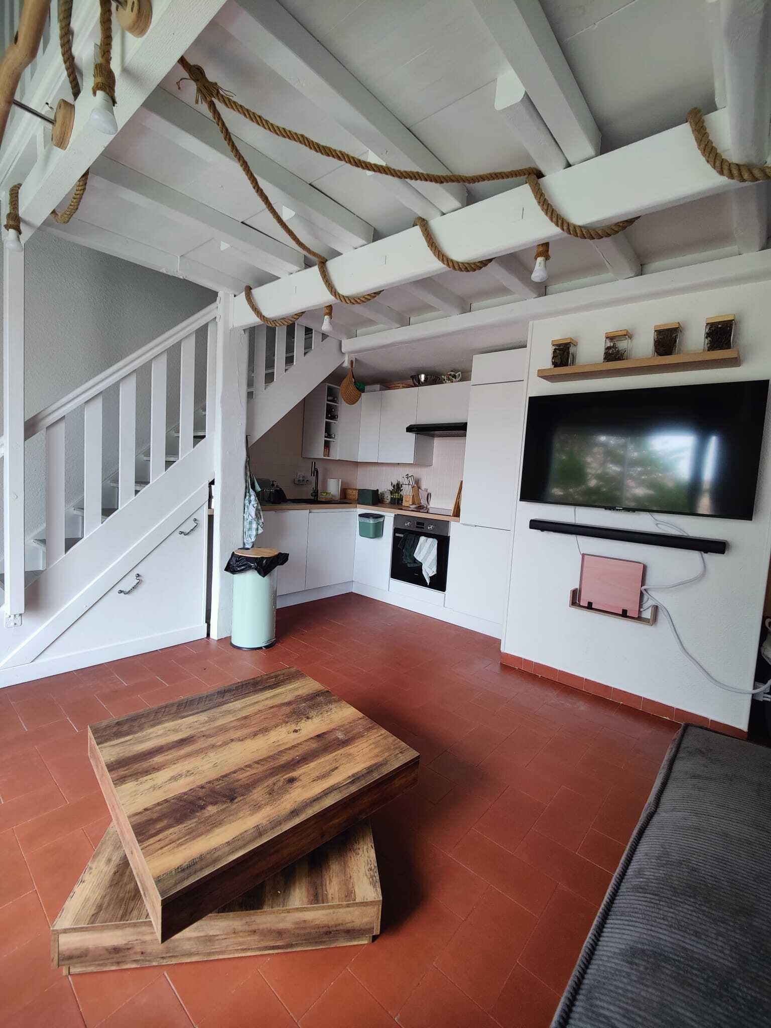 A compact living space with a wooden table, a TV mounted on the wall, and a staircase leading to an upper level.