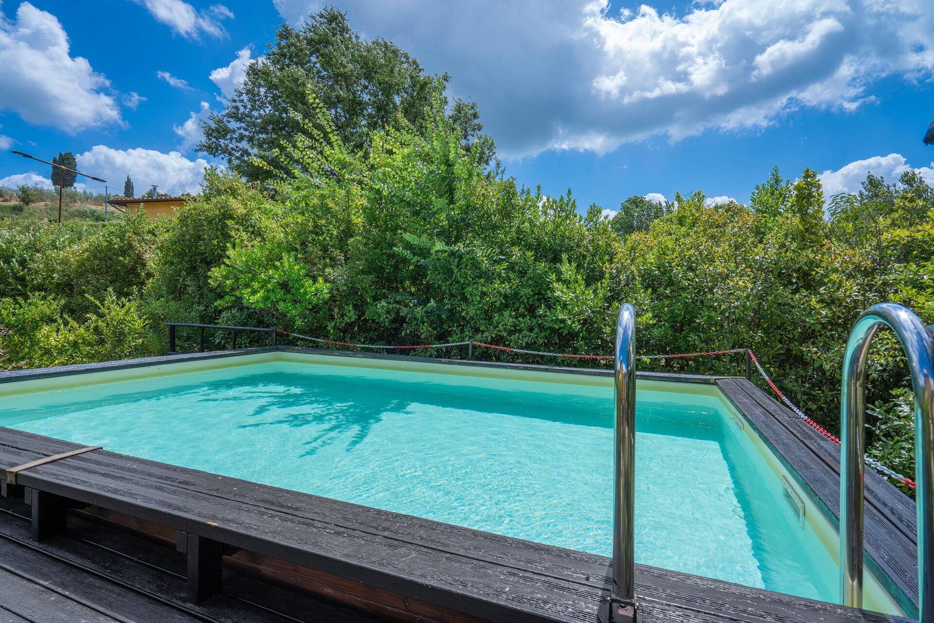 Ein rechteckiger Swimmingpool mit einer Holzterrasse im Vordergrund, umgeben von üppigem Grün und Bäumen.