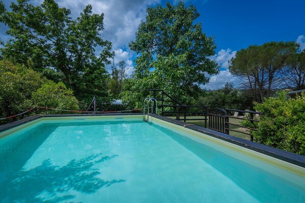 A rectangular swimming pool with a wooden deck, surrounded by lush greenery and trees.