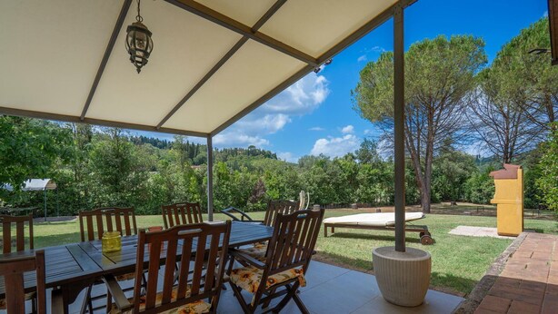 En udendørs patio med et bord og stole, en pergola samt udsigt over et skovområde.