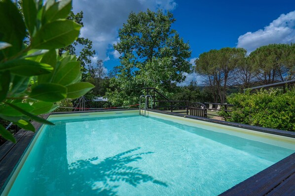 A rectangular swimming pool with a wooden deck surrounding it, set in a lush, green outdoor area with trees and shrubs.