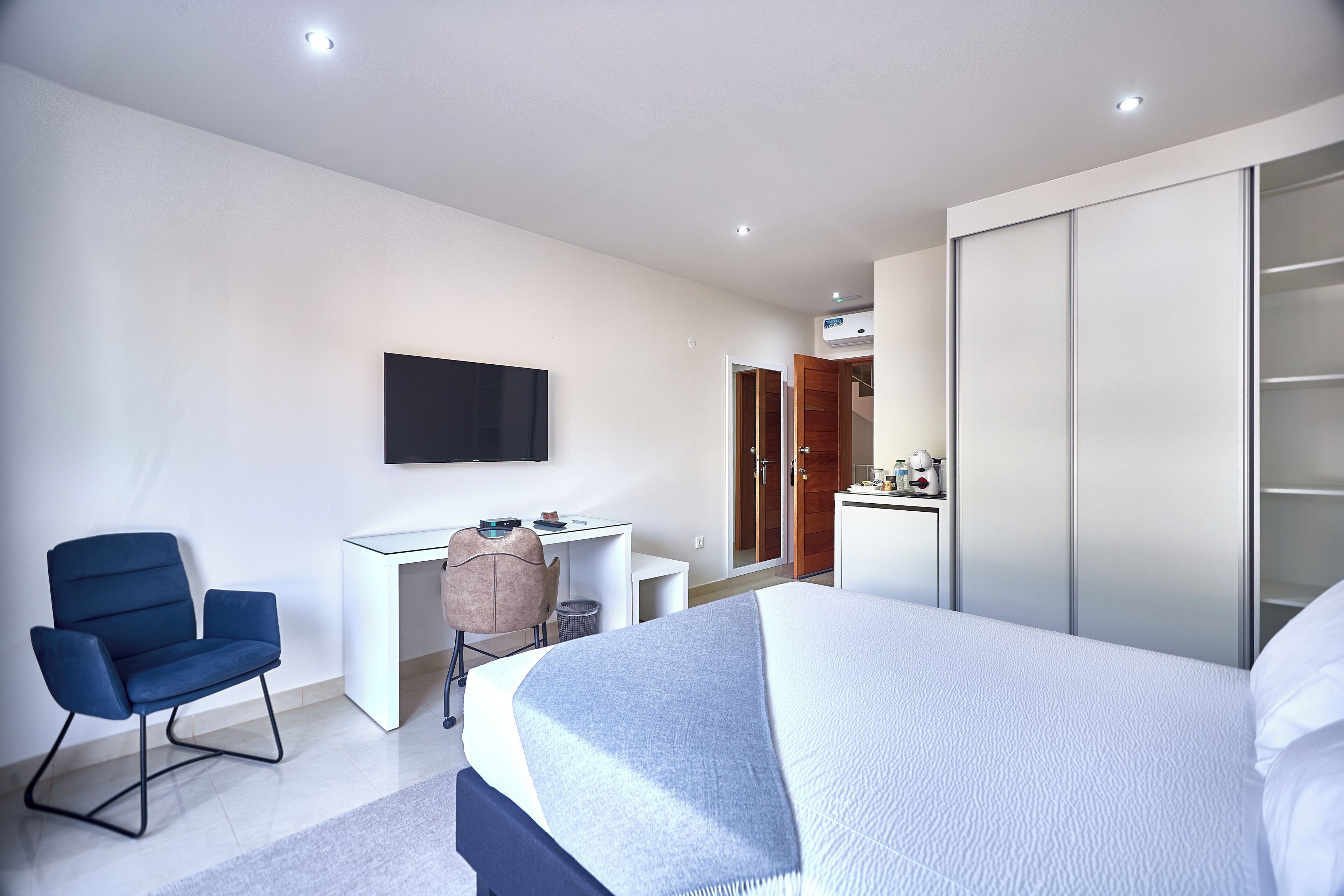 A modern hotel room with a large bed, a desk, a chair, a TV mounted on the wall, and a wardrobe.
