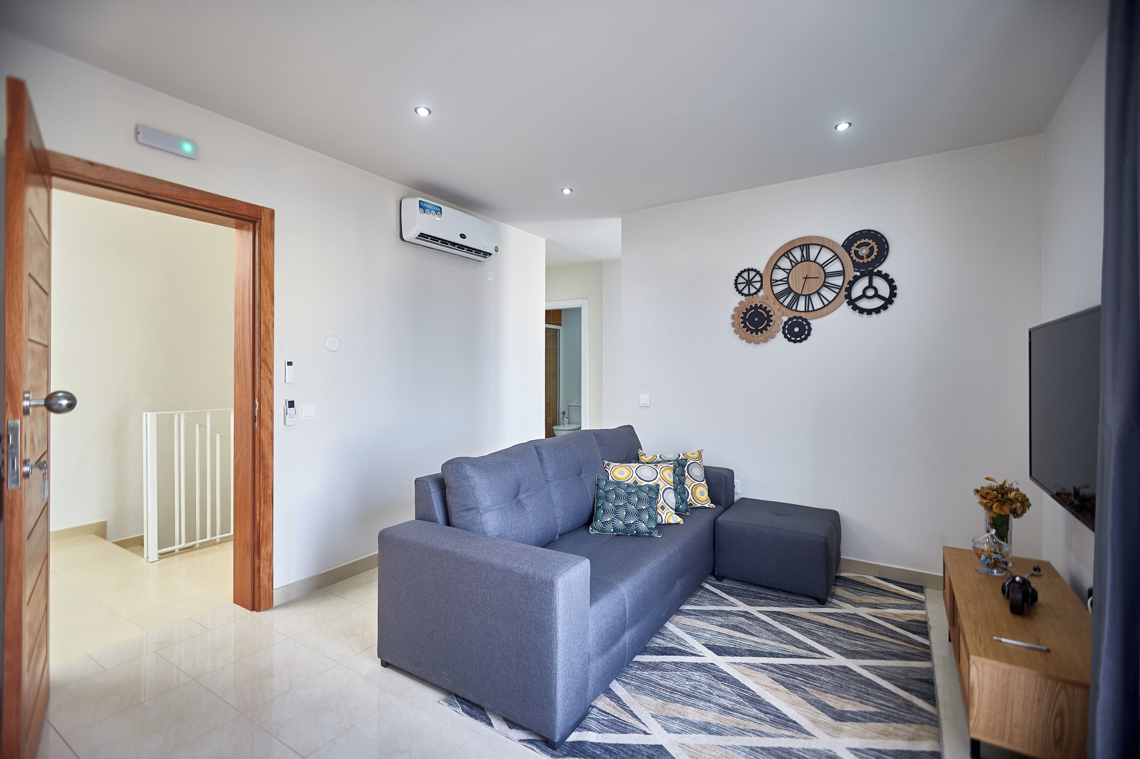 Una sala de estar moderna con un sofá gris, una alfombra estampada y un reloj montado en la pared.