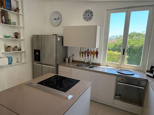 A modern kitchen with a built-in stove, refrigerator, and oven. There are decorative plates on the wall, a window with a view of greenery, and a shelf with kitchen utensils.