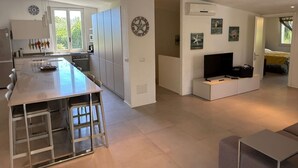 A modern kitchen with a central island, a dining area with a bar stool, a living room with a TV and seating, and a bedroom visible through an open door. - Beautiful private villa for 11 guests with WIFI, A/C, TV, patio and panoramic view (Villasimius)