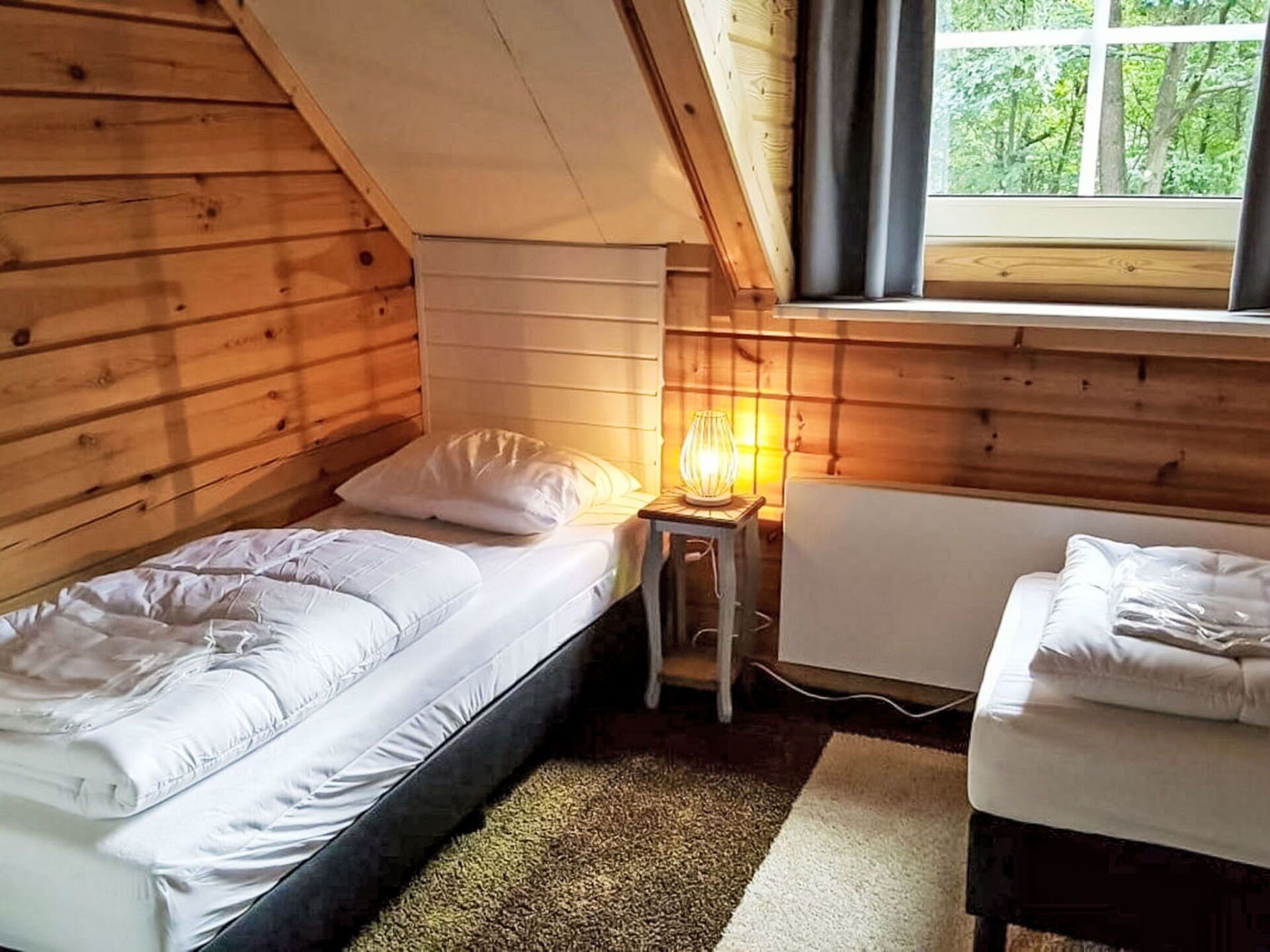 A small, cozy room with wooden walls and a sloped ceiling. There are two beds with white bedding, a small table with a lamp, and a window with a view of trees.