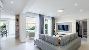 Un salon moderne avec un canapé d’angle gris, un téléviseur à écran plat fixé au mur et un coin repas comprenant une table et des chaises.