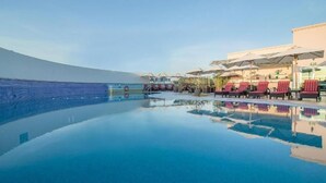Une magnifique piscine à débordement d’un bleu profond, agrémentée de chaises longues et de parasols, et entourée d’un bâtiment doté d’un toit incurvé.