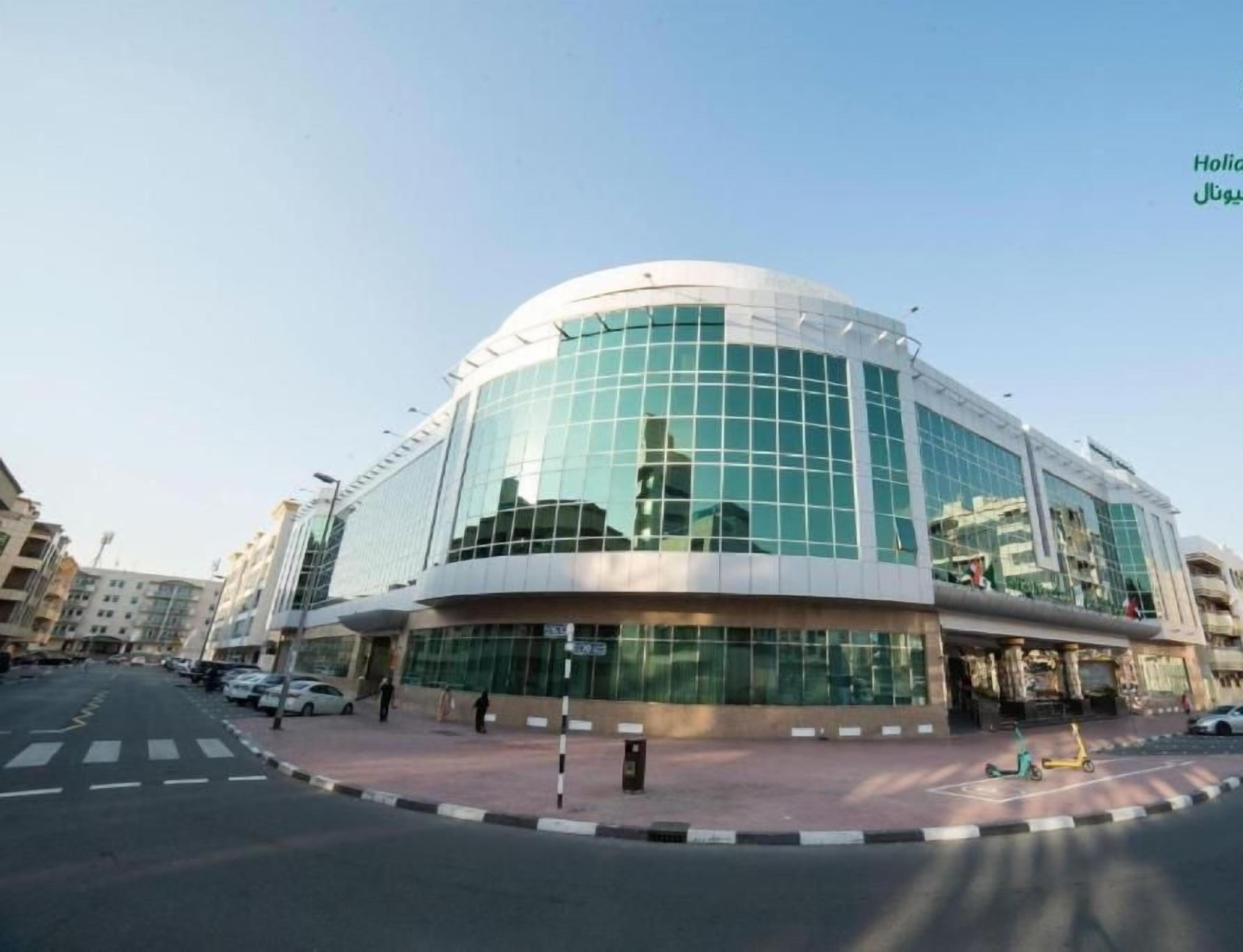 A modern building with a curved glass facade, a pedestrian area with a yellow scooter, and parked cars.