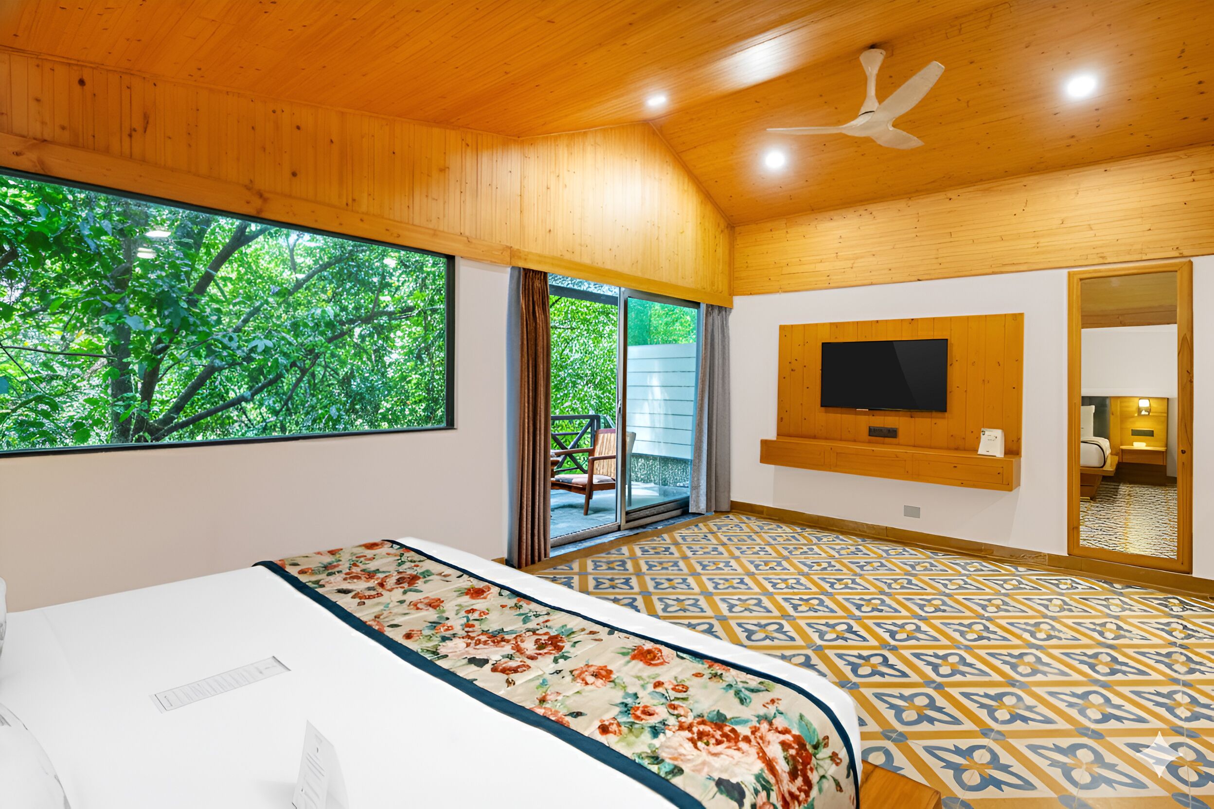 Een slaapkamer met een groot raam, een bed met een bloemenprint, een plafondventilator en een aan de muur bevestigde televisie.