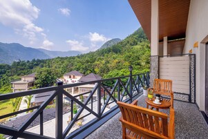 A balcony with a wooden chair and table, overlooking a lush green valley and distant mountains.