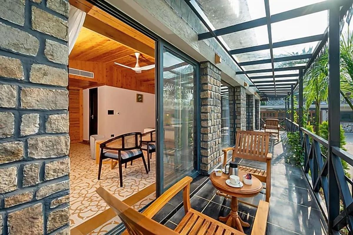 A cozy indoor space with a wooden ceiling, stone walls, and a glass door leading outside. There are wooden chairs and a small round table with a cup and saucer.