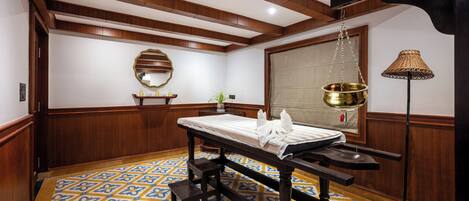 A room with a wooden ceiling, a patterned floor, a bed with a white mattress, a bedside table, a mirror, and a hanging lamp.