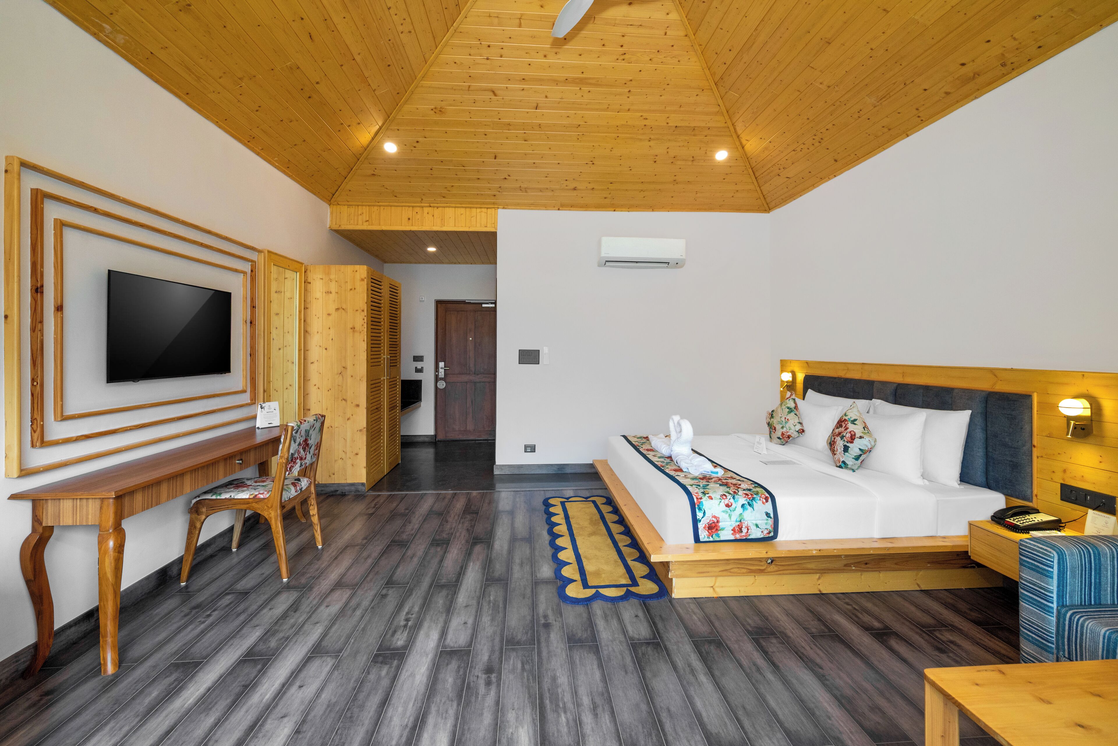 A hotel room with a wooden ceiling, a bed with floral bedding, a TV mounted on the wall, a wooden desk, and a chair.