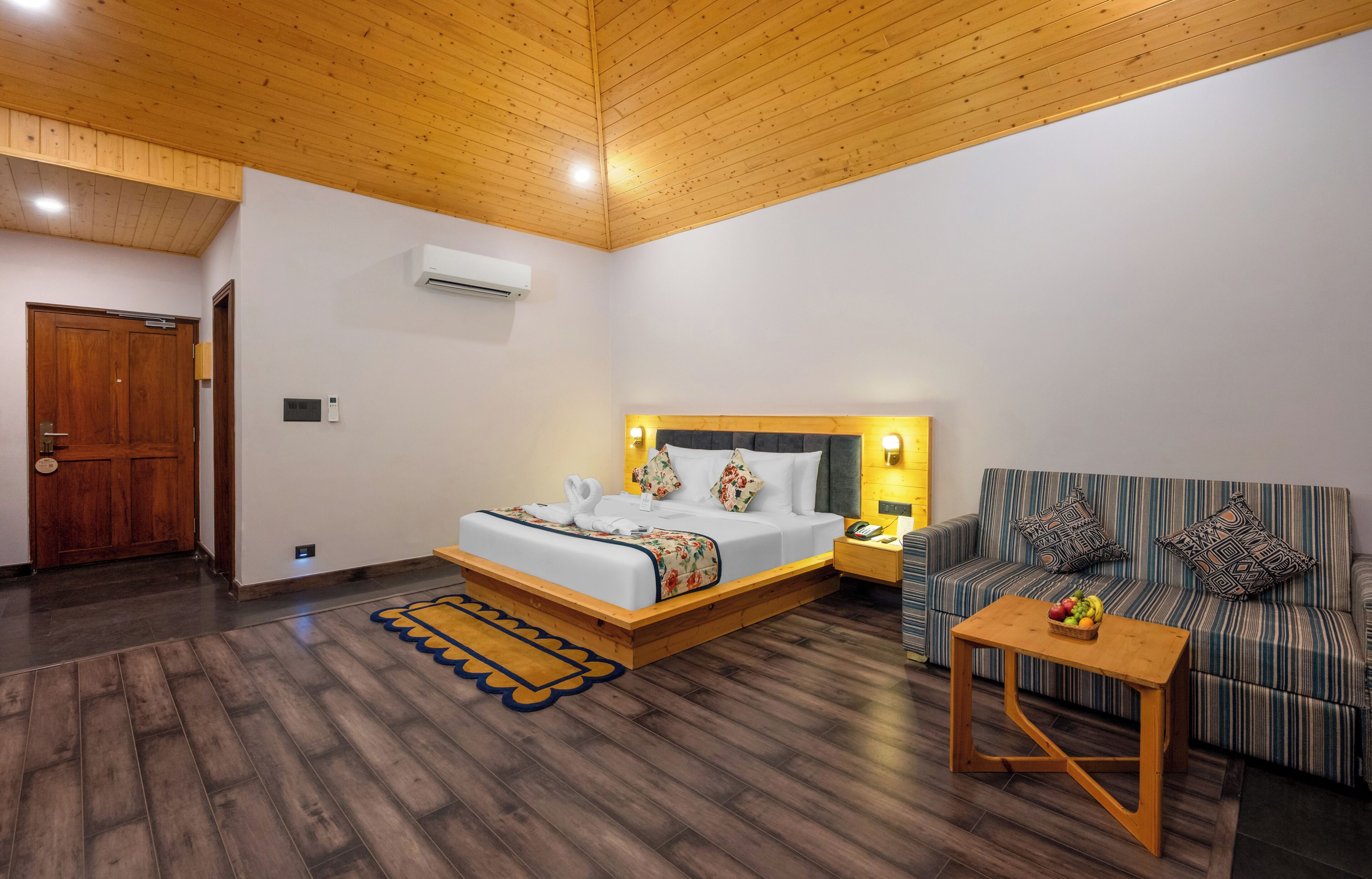 A hotel room with a wooden ceiling, a bed with a headboard, a sofa, a small table, and a wooden floor.