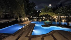 A nighttime swimming pool area with multiple pools, palm trees, and a water tower in the background. - Jewel's Homey Place -2br in a Resort-like Condo (Pasig)