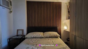 A bedroom with a wooden headboard, a bedside table with a lamp, and a bed with floral bedding. - Jewel's Homey Place -2br in a Resort-like Condo (Pasig)