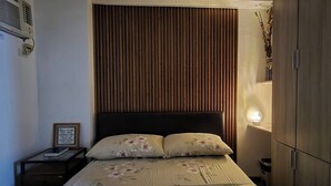 A bedroom with a wooden headboard, a bedside table with a lamp, and a bed with floral bedding. - Jewel's Homey Place -2br in a Resort-like Condo (Pasig)