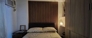 A bedroom with a wooden headboard, a bedside table with a lamp, and a bed with floral bedding. - Jewel's Homey Place -2br in a Resort-like Condo (Pasig)