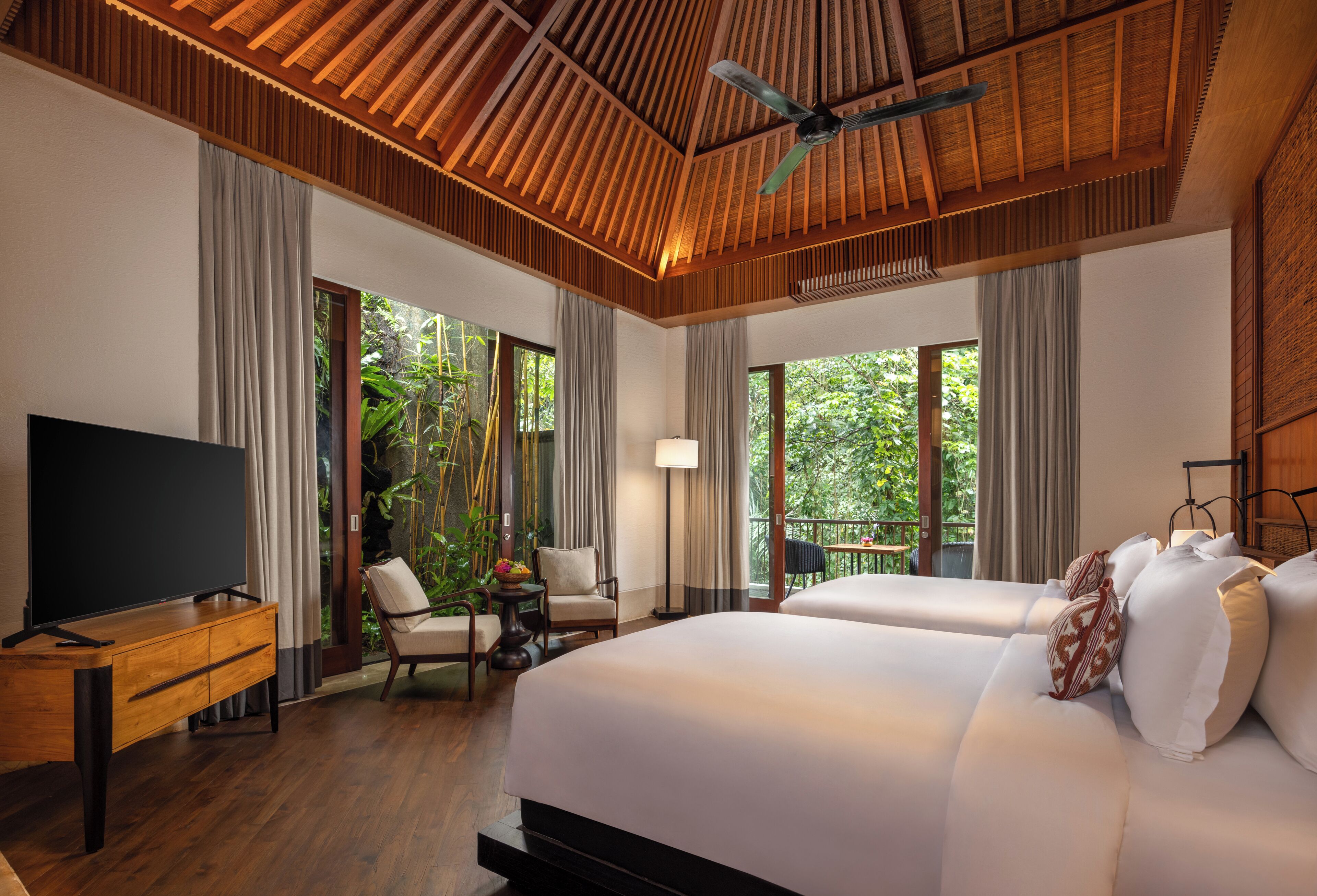 A hotel room with a large bed, a TV, a desk, and a balcony with a view of greenery.