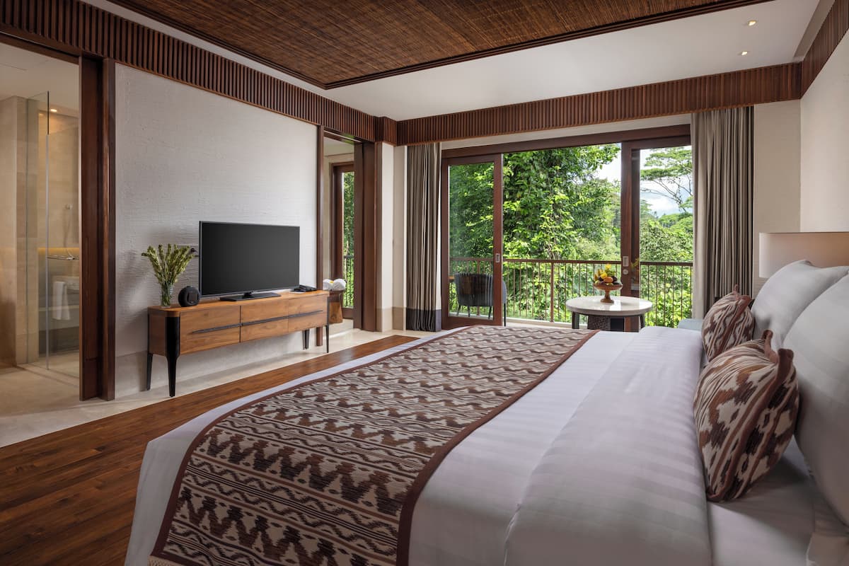 Una habitación de hotel moderna con una cama grande, un televisor sobre una consola de madera, un balcón con vistas a la vegetación y una pequeña mesa redonda.