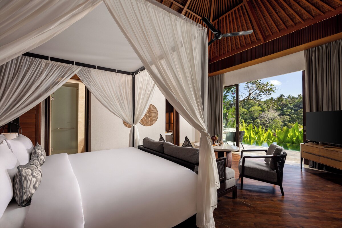 Quarto com cama grande, televisão, área de refeições com mesa e cadeiras, e vista para o verde lá fora.