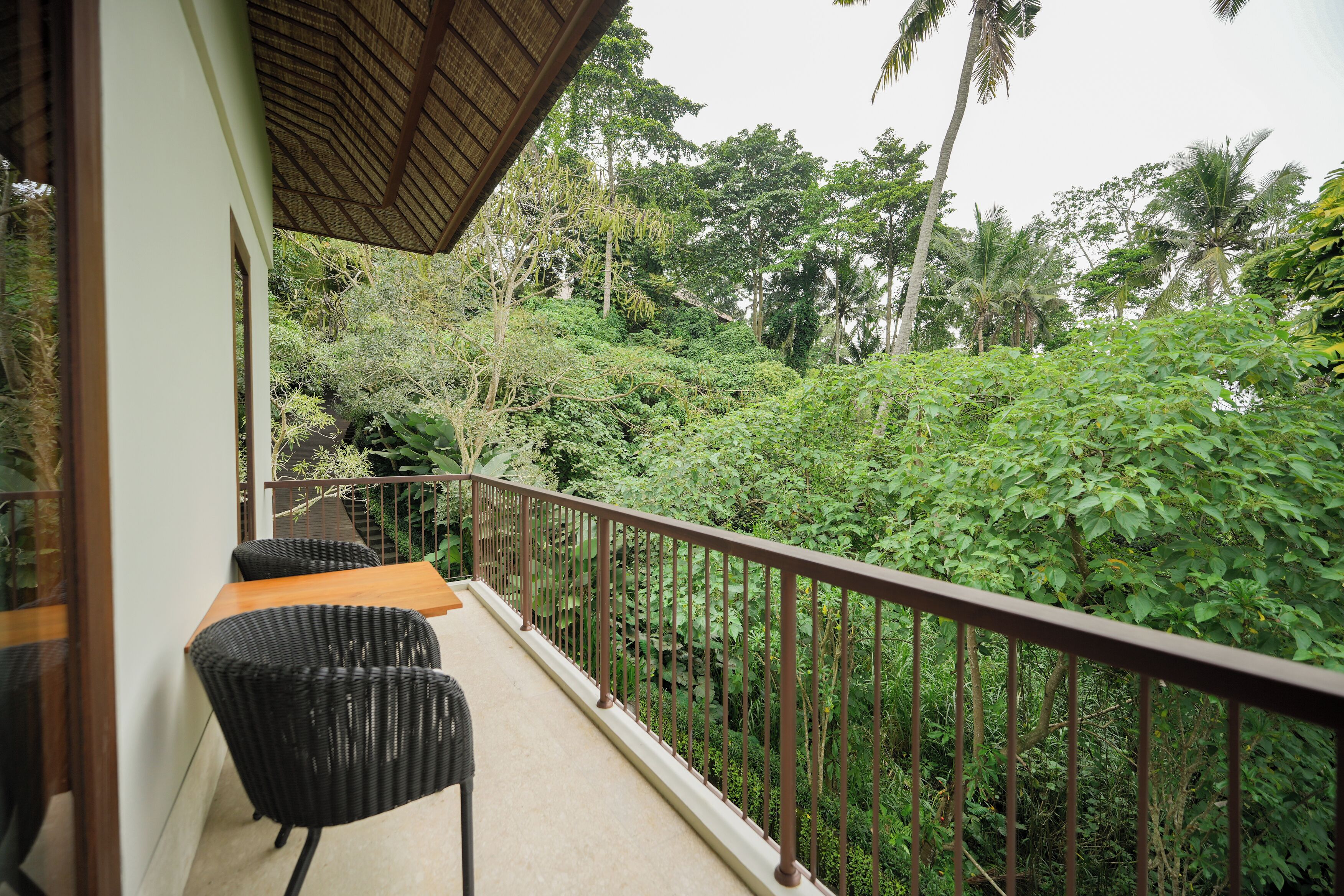 A balcony with wicker chairs, a wooden table, and a view of dense tropical foliage.