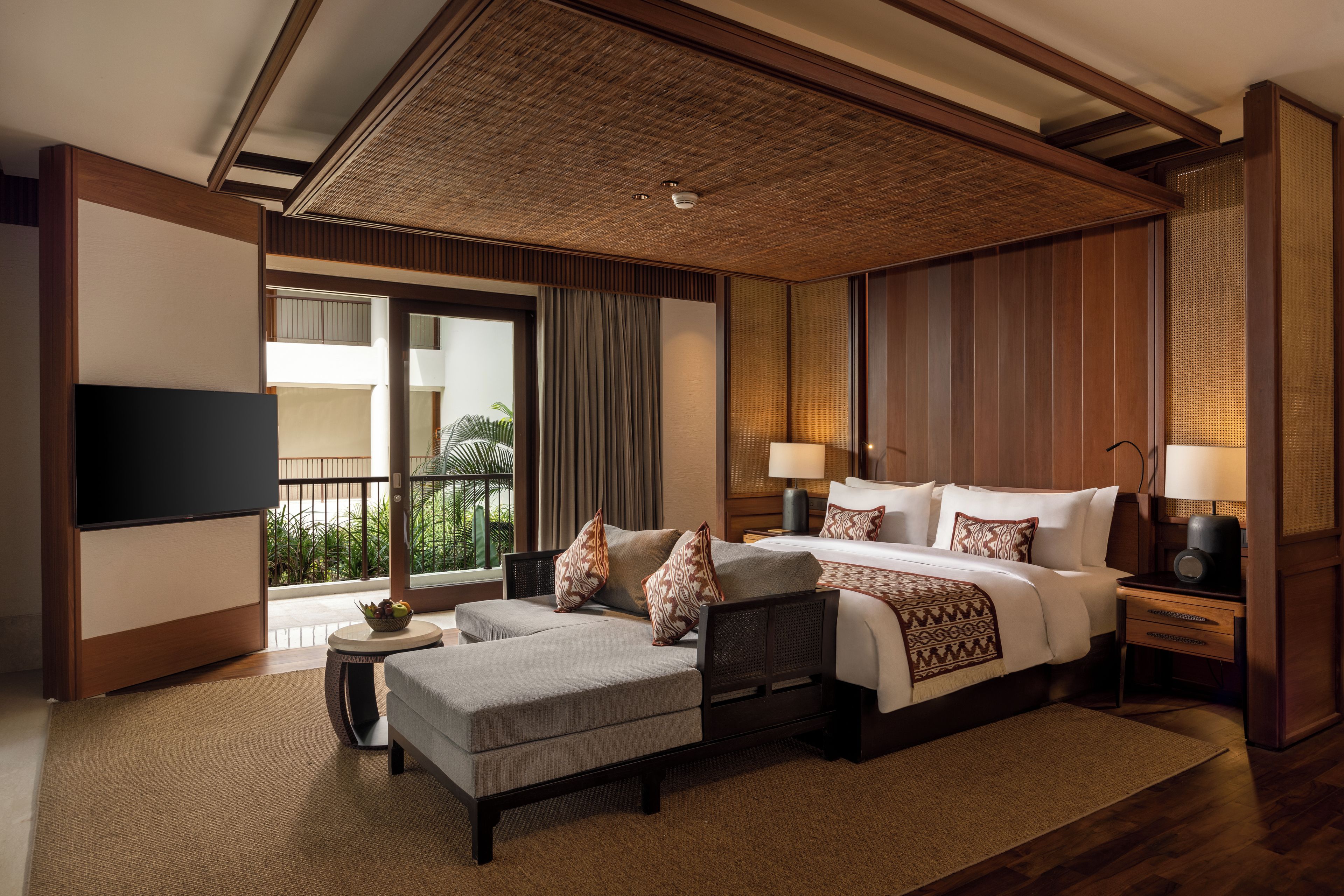 A hotel room with a bed, a sofa, a TV, a balcony, and a view of greenery.