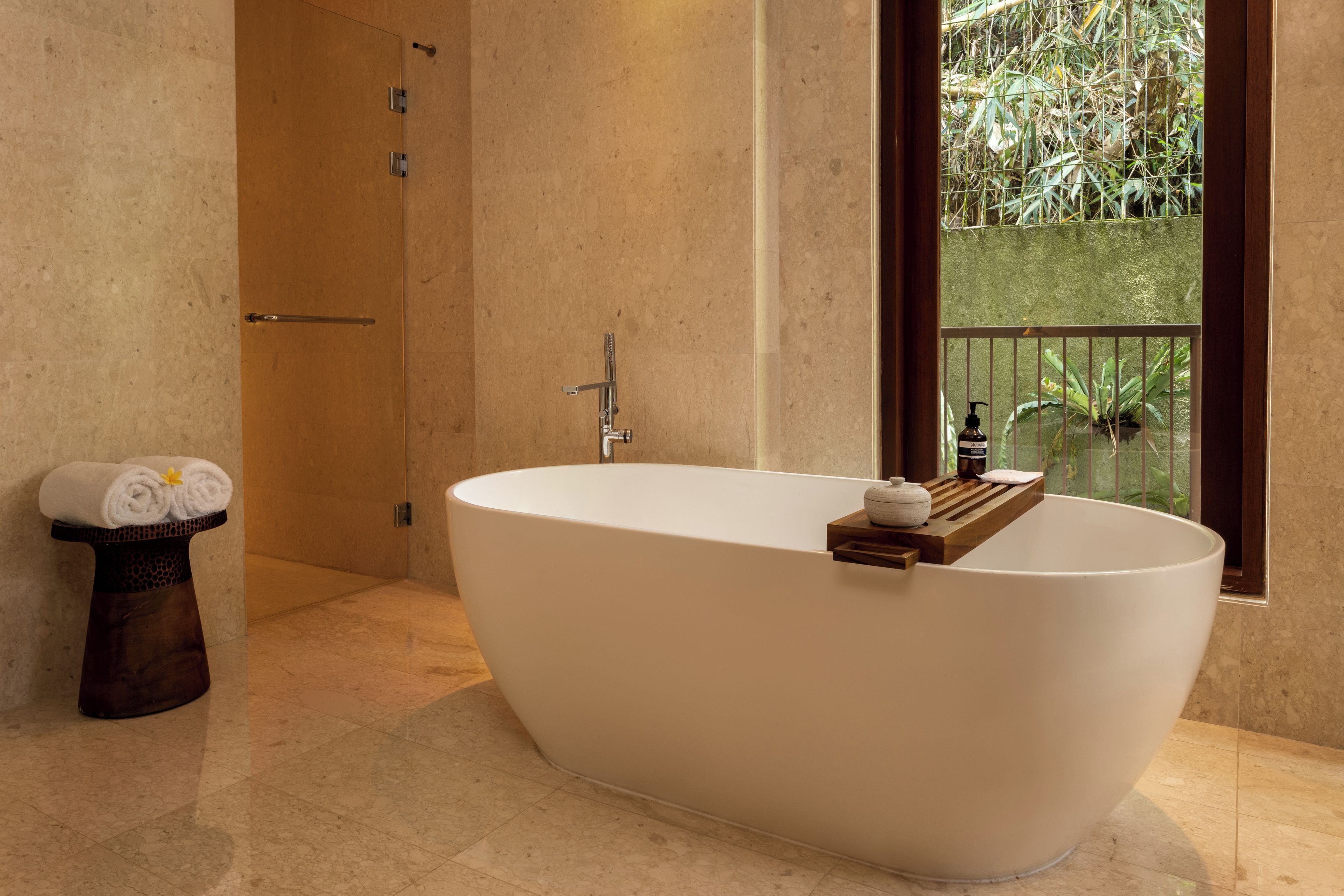 A modern bathroom with a freestanding bathtub, a wooden towel rack, and a view of greenery through a window.