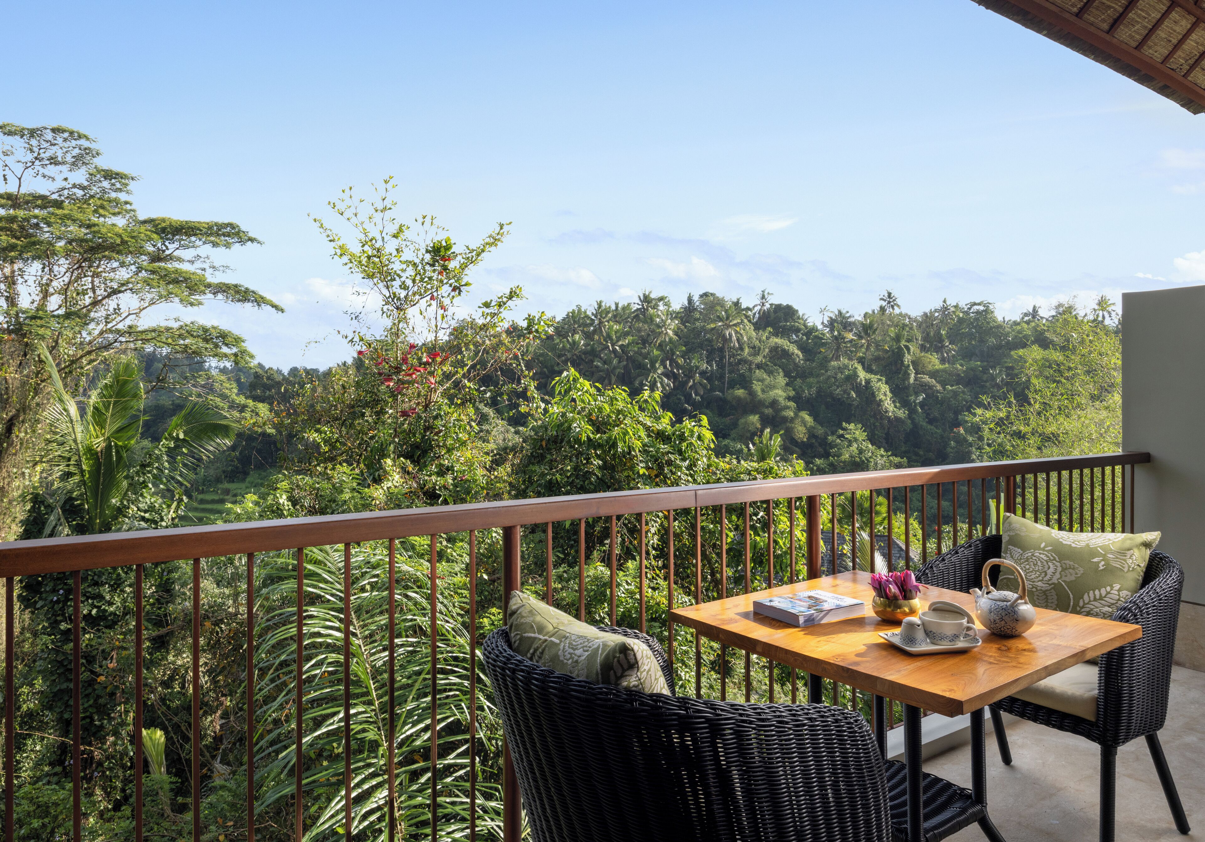A balcony with wicker furniture, a table with a teapot, and a view of a lush forest.