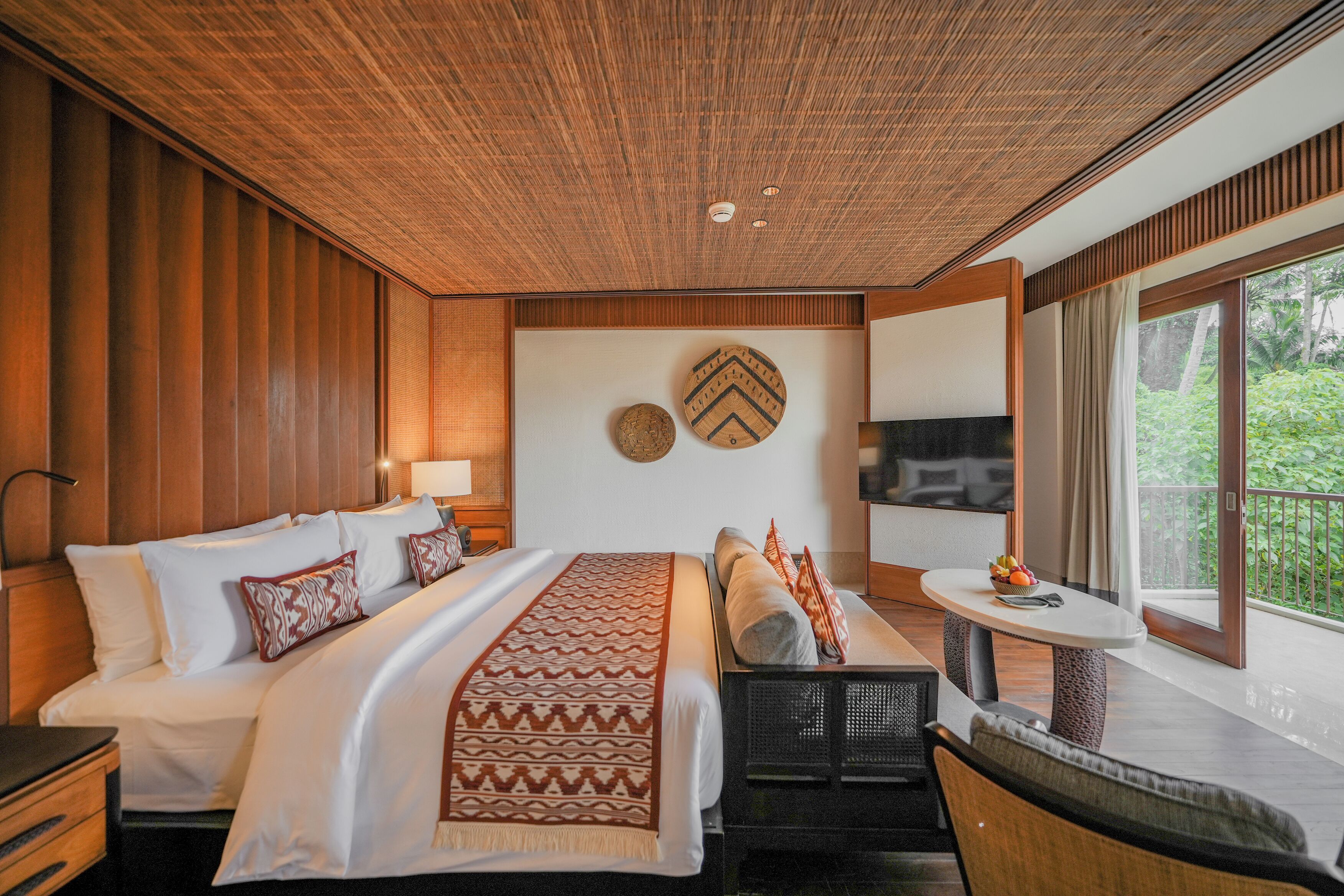 A modern hotel room with a wooden ceiling, a large bed with white linens, a sofa, a small table, and a balcony with a view of greenery.