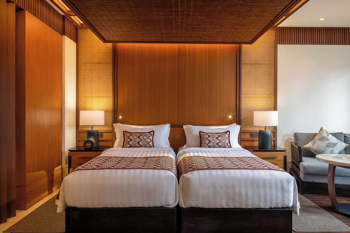 Una cama doble con colcha estampada, dos mesitas de noche con l ámparas, un cabecero de madera y una zona de estar con una silla y una mesa.